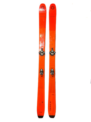 Two orange skis with bindings on a gray carpeted floor.