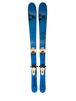 A pair of blue Fischer skis with white bindings, standing vertically against a light wood-paneled wall next to a window.