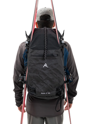 Person wearing a black backpack with a brand logo, holding skis over their shoulder on a white background