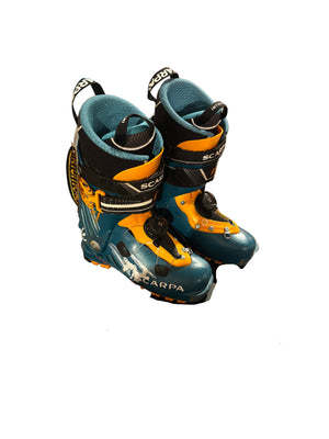 Pair of blue and yellow ski boots on a black box with visible branding in an indoor setting.