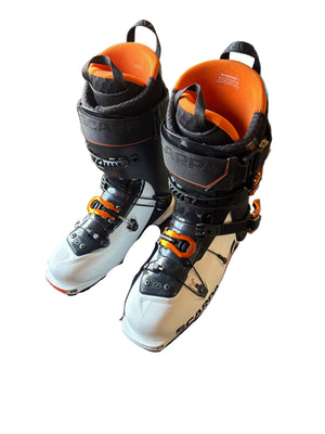 Pair of black and white ski boots with orange accents in a box on a wooden floor.