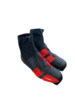A pair of black and red ski boots resting on a cardboard box in an office or workshop setting. The box has a visible Vacuvm logo, and other office items are visible in the background.