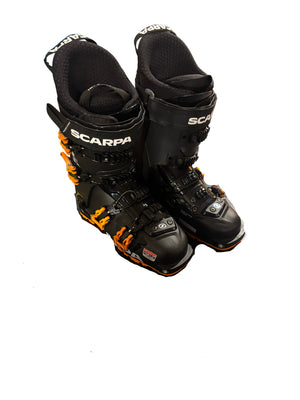 Black ski boots with orange accents on a black mat in an indoor setting.